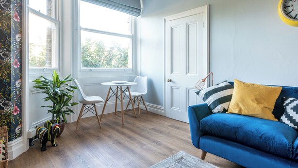 Ein Zimmer mit einem blauen Sofa, einem gelben Kissen, einer weißen Tür und einem Fenster mit Blick auf Bäume.