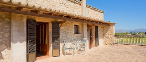 Un edificio de piedra con un patio empedrado, contraventanas de madera y una mesa con sillas.