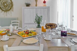 A breakfast table set with fruits, pastries, and beverages, with a white cabinet and a plant in the background.