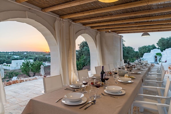 A long dining table set for a meal with white chairs, set against a backdrop of a sunset and a view of the countryside.