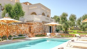 Ein Poolbereich mit Liegestühlen und Sonnenschirmen, eine Steinmauer und ein Gebäude mit Bögen im Hintergrund.
