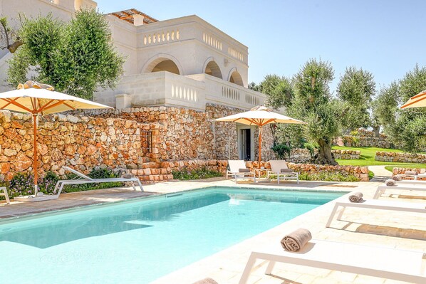 A pool area with lounge chairs and umbrellas, a stone wall, and a building with arches in the background.