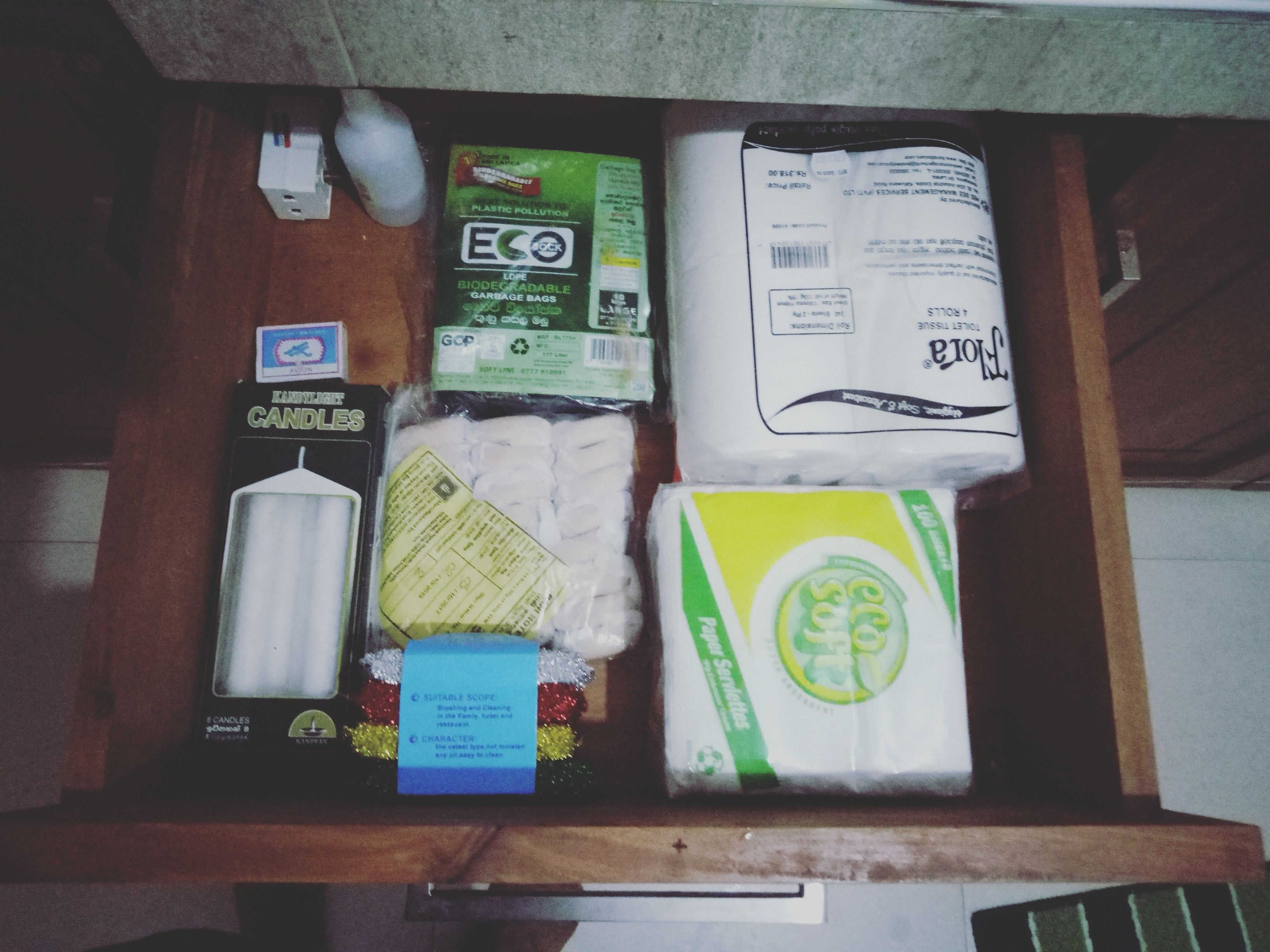 Un cajón con varios objetos, incluyendo una vela, un paquete de papel higiénico, una caja de pañuelos y un paquete de toallitas.