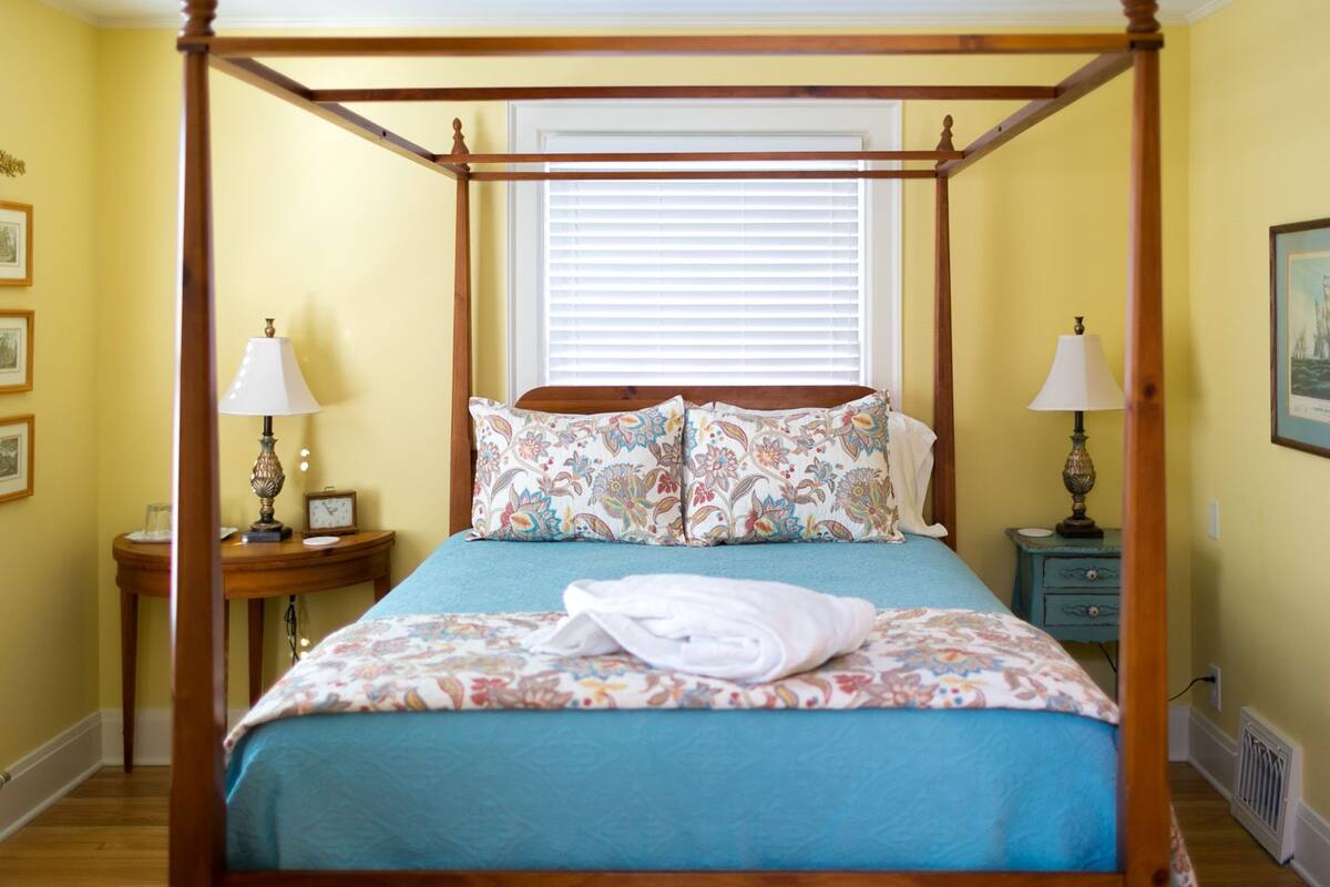 A four-poster bed with floral bedding, two bedside tables with lamps, and a window with blinds.