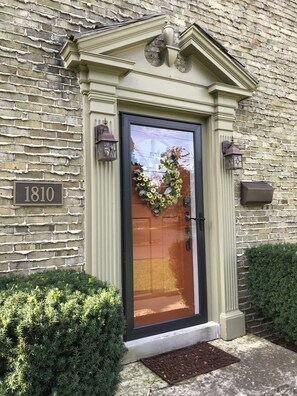 Um edifício com fachada de tijolos, uma porta decorada com uma coroa de flores e uma placa com o número 1810.