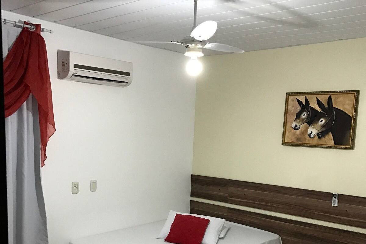 A single-bed room with a ceiling fan, an air conditioning unit, a red curtain, a wall-mounted painting of donkeys, and a bedside table.