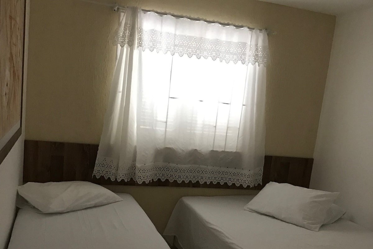 A room with two single beds, a window with a white lace curtain, and a wooden headboard.