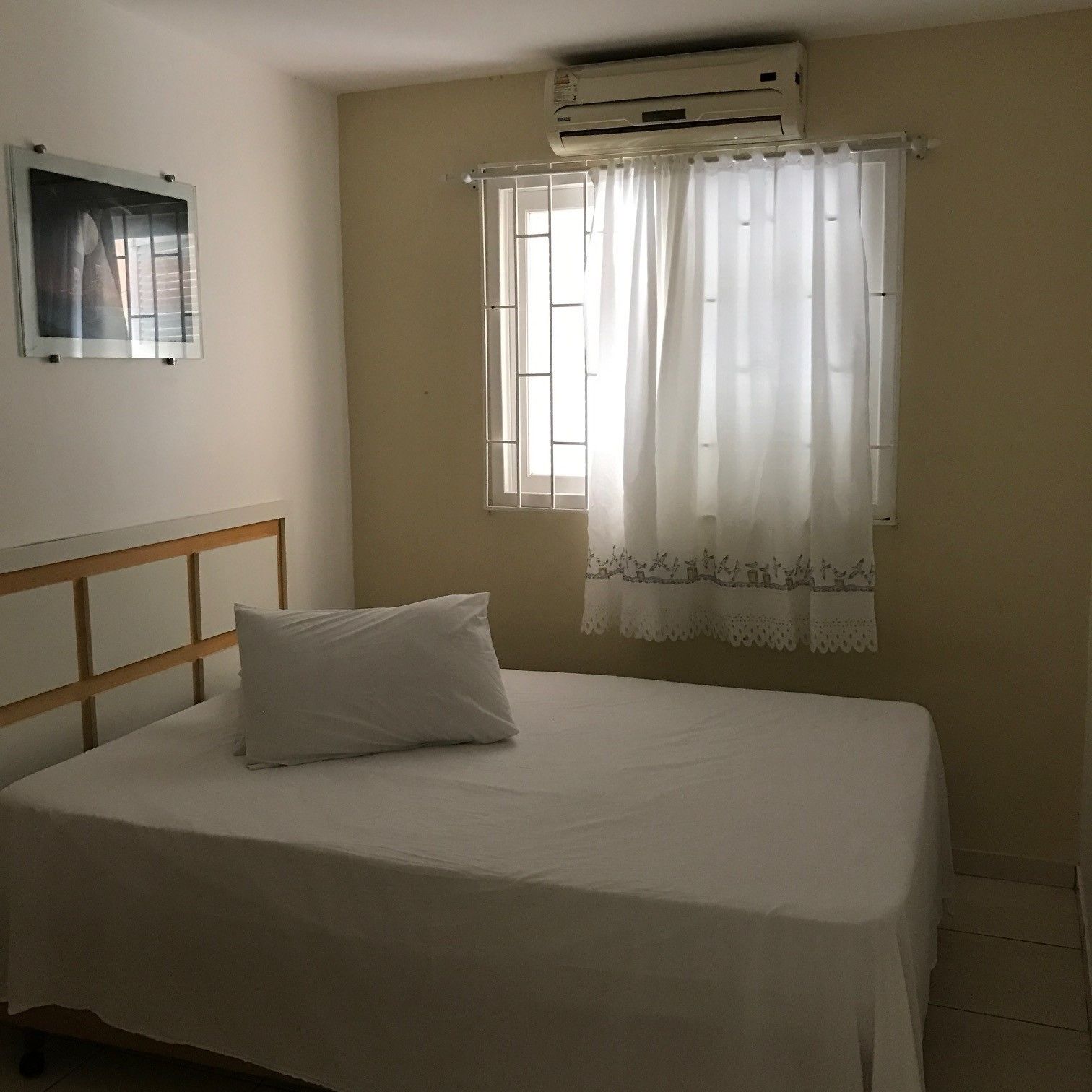 A bedroom with a bed, a window with curtains, an air conditioning unit, and a wall-mounted picture.
