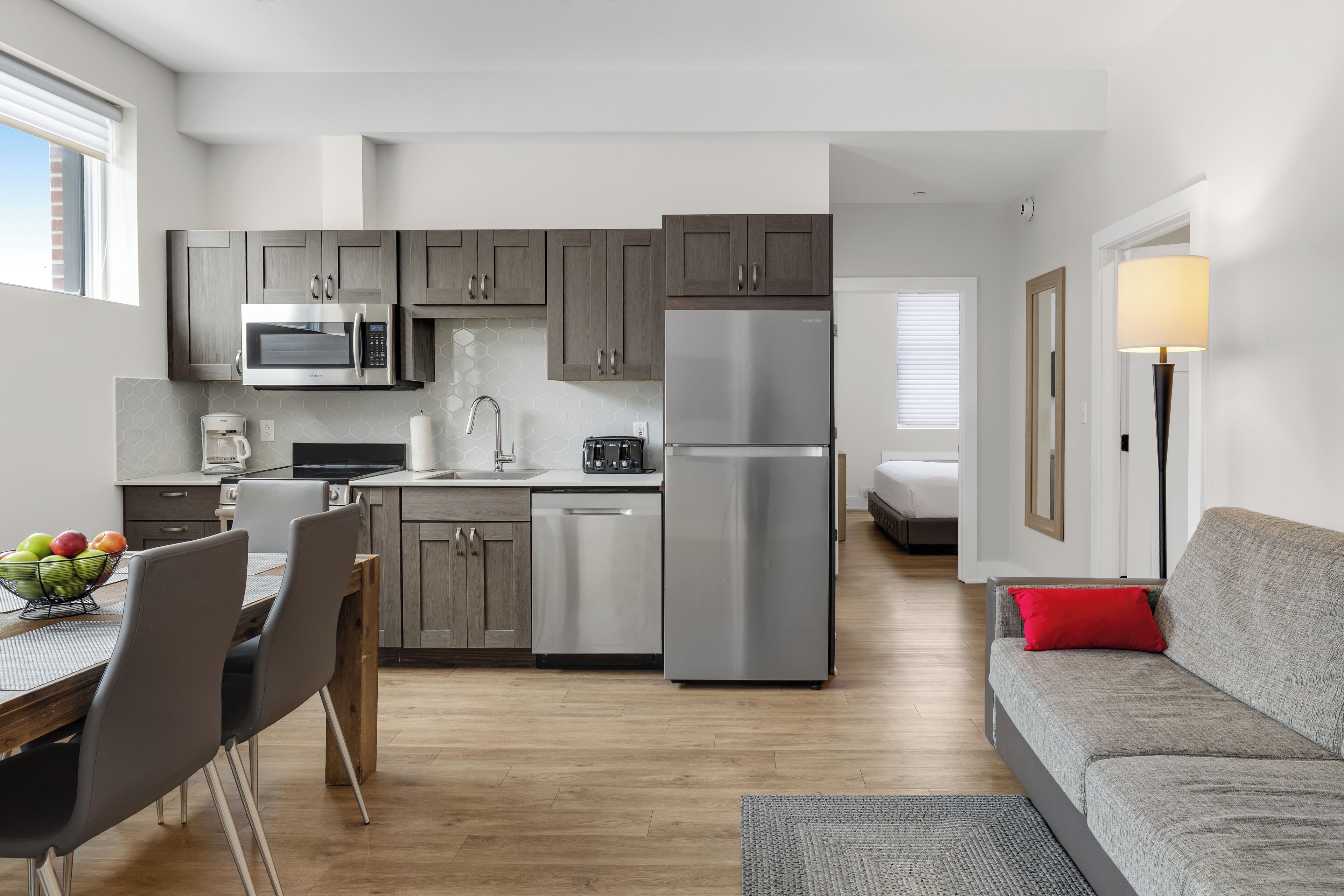 A modern kitchen with stainless steel appliances, a dining area with a wooden table and chairs, and a grey sofa with a red pillow.