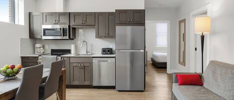 Una cocina moderna con electrodomésticos de acero inoxidable, un comedor con mesa y sillas de madera, y un sofá gris con un cojín rojo.
