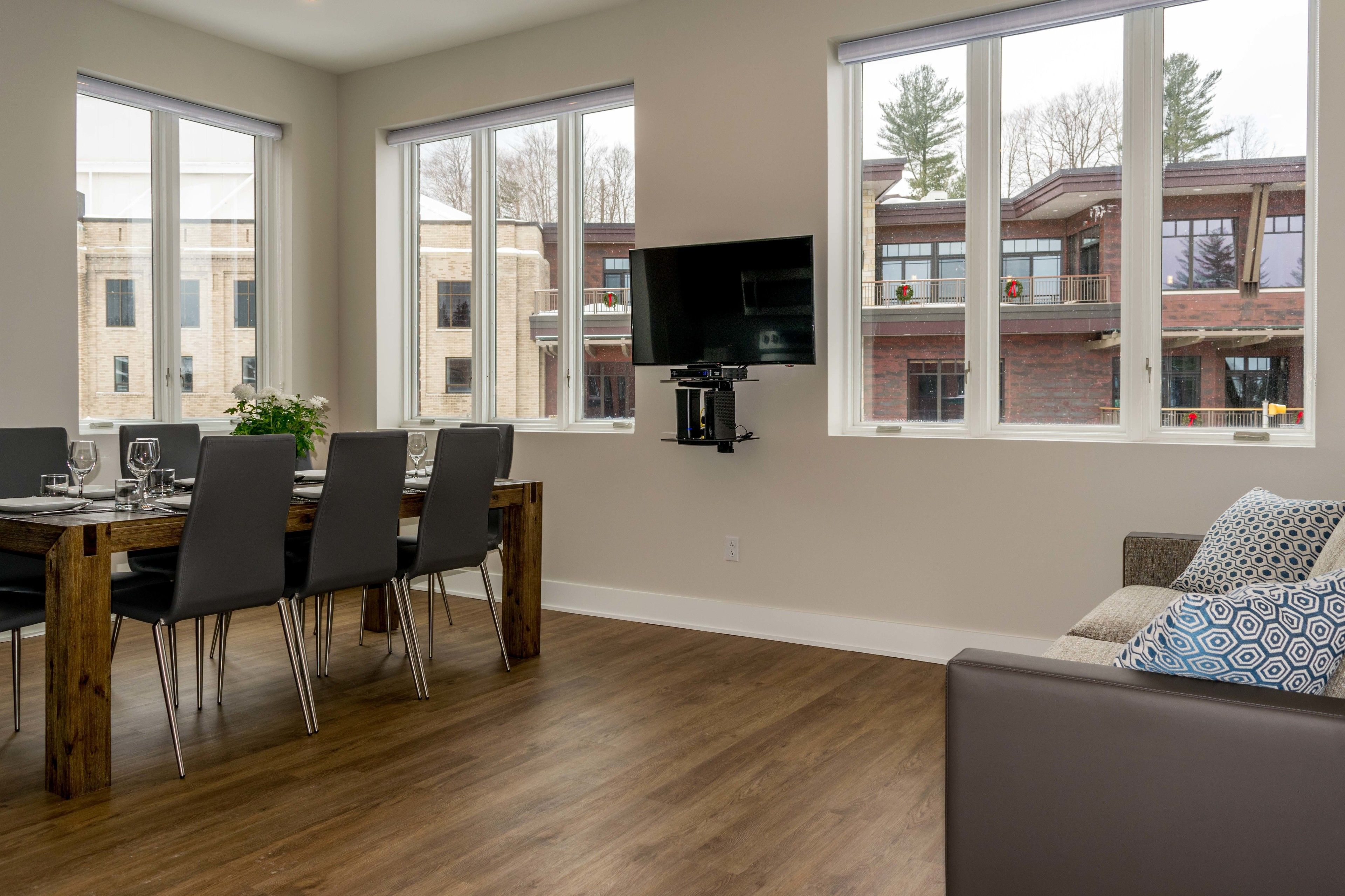 Un comedor con una mesa y sillas de madera, un televisor de pantalla plana montado en la pared y ventanas grandes con vistas a un edificio con balcones.