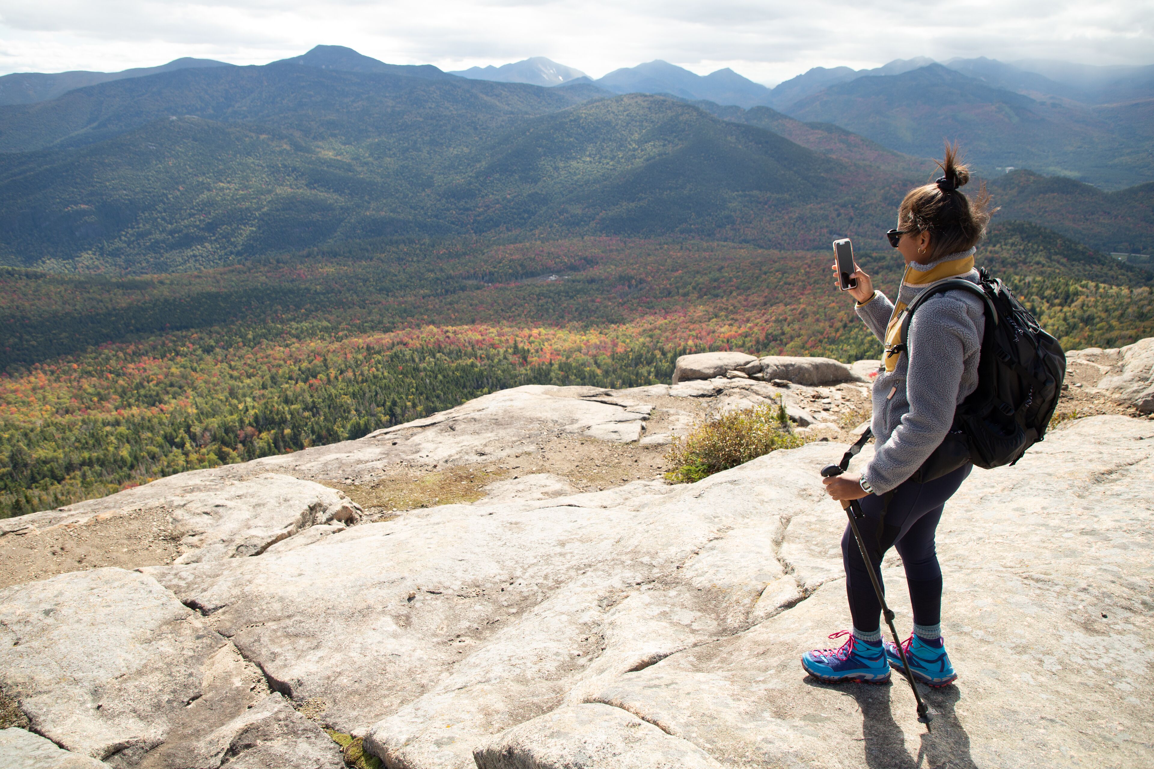 A person standing on a rocky outcrop with a smartphone, overlooking a mountainous landscape.