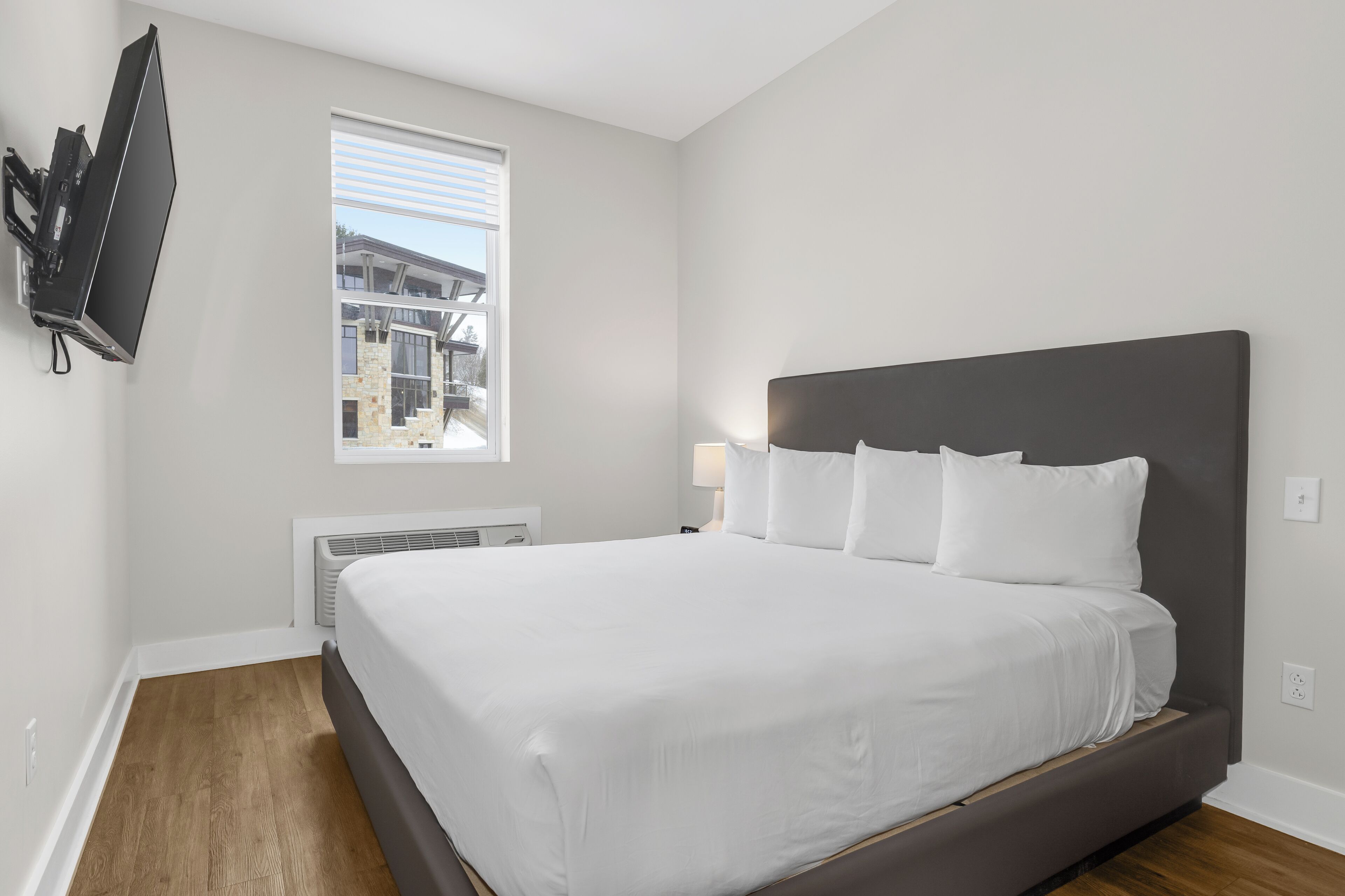 A modern hotel room with a large bed, a flat-screen TV mounted on the wall, and a window with blinds.