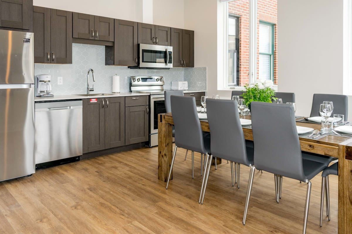 Una cocina moderna con armarios oscuros, electrodomésticos de acero inoxidable y una zona de comedor con mesa de madera y sillas grises.