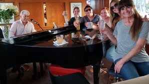 A group of people seated around a piano in a room with wooden paneling and a clock on the wall.