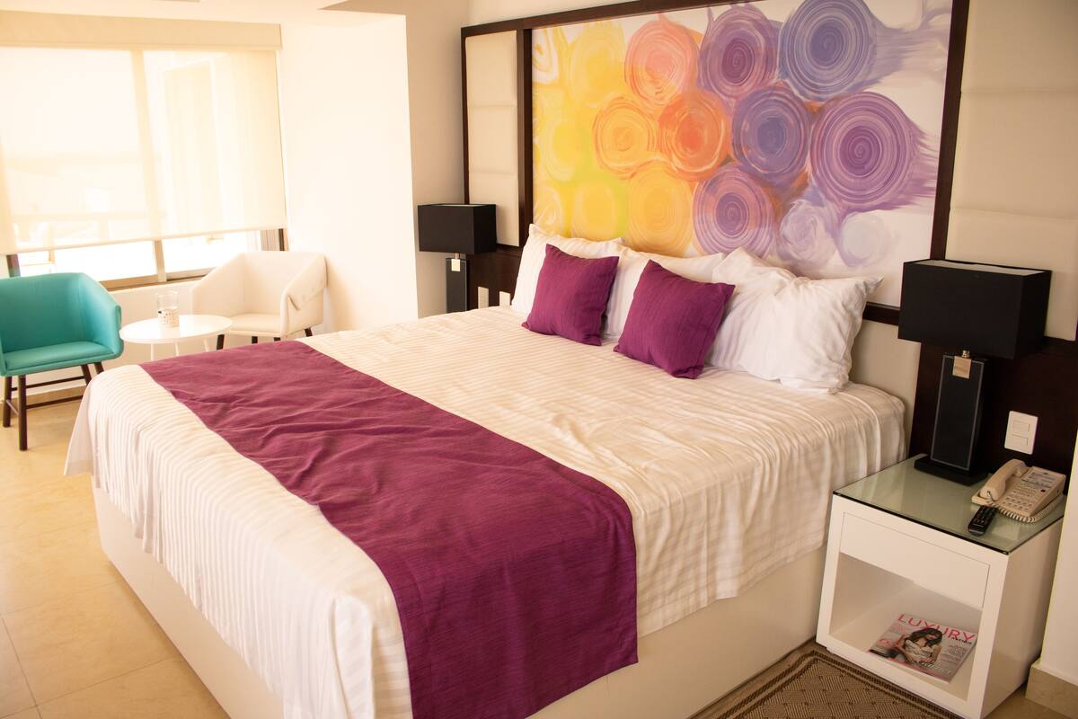 A hotel room with a large bed, two bedside tables, a nightstand, a chair, and a window with blinds.
