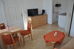 Ein Zimmer mit Holztisch, zwei Stühlen, einem Fernseher auf einem Schrank, einer kleinen Küchenzeile und einer Tür.