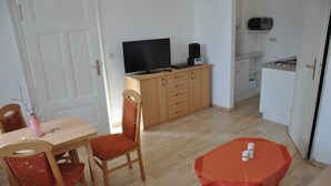 A room with a wooden dining table, two chairs, a television on a cabinet, a small kitchen area, and a door. - Apartment Gording II - Holiday residence sea desire (Koserow)