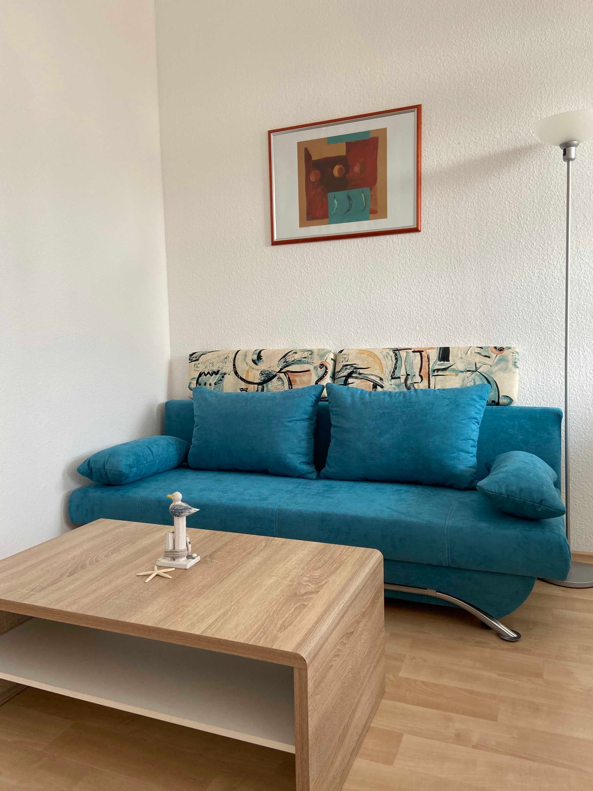 Un sofá azul verdoso con un cuadro abstracto enmarcado encima, una mesa de centro de madera con un objeto decorativo, y una lámpara de pie a la derecha.