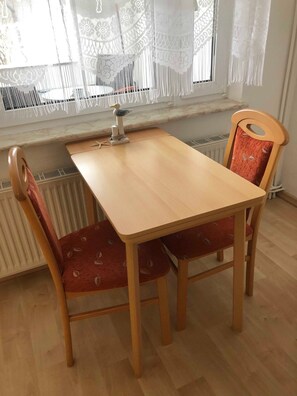 Een houten tafel met twee stoelen, een decoratief beeld van een vogel, kantgordijnen en een radiator.