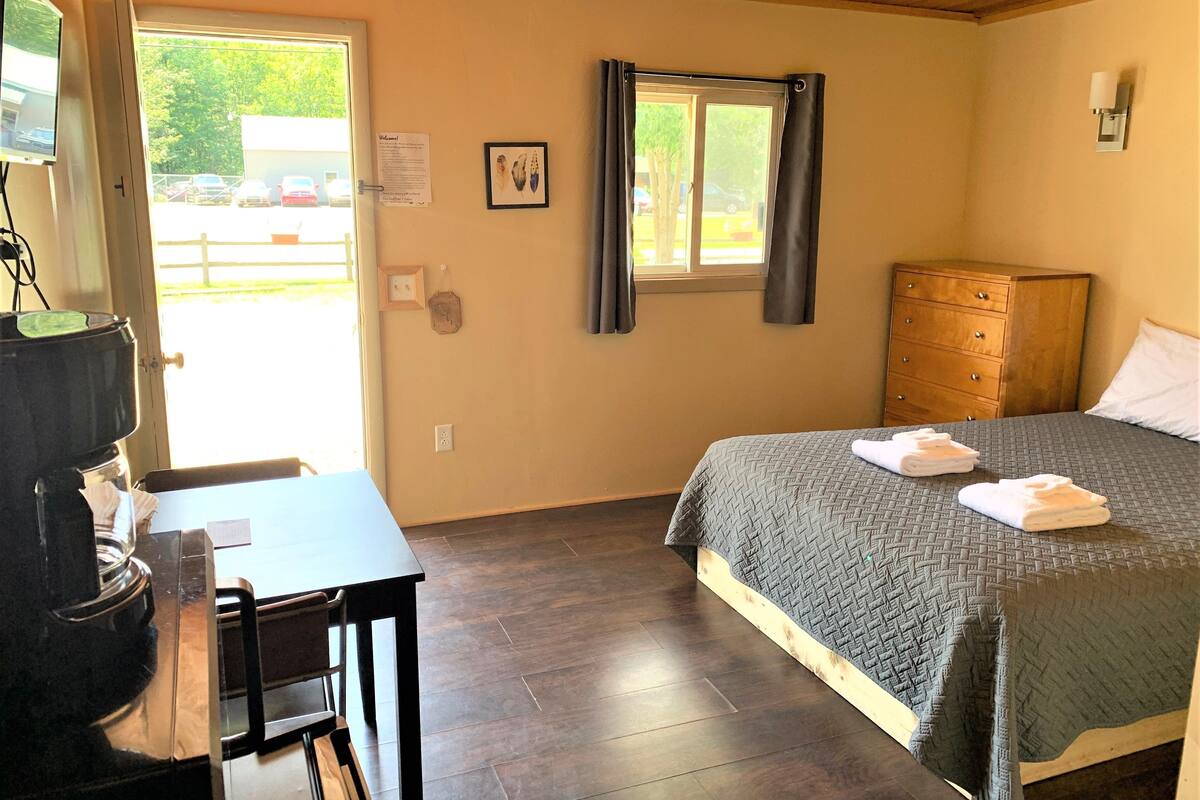 A hotel room with a bed, a wooden dresser, a desk with a chair, a coffee machine, a window with curtains, and a view of the outside parking area.