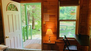 A wooden cabin room with a bed, a desk, a chair, a window with a view of greenery, and a door leading outside. - Pine Crest Motel And Cabins (Barton)
