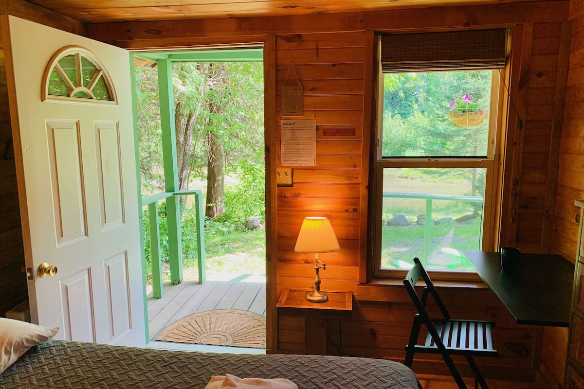 A wooden cabin room with a bed, a desk, a chair, a window with a view of greenery, and a door leading outside.