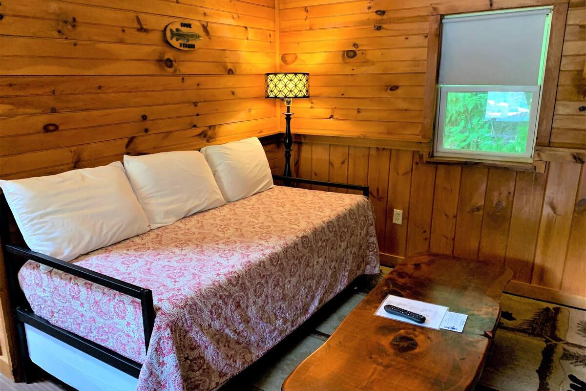 A wooden cabin room with a bed, a table, a lamp, and a window with a view of greenery.