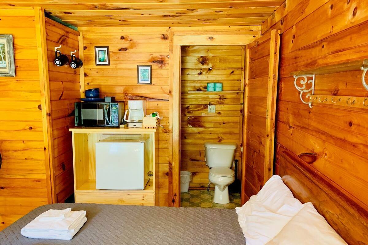 A wooden cabin room with a bed, a small kitchenette with a microwave, coffee maker, and refrigerator, and a toilet.