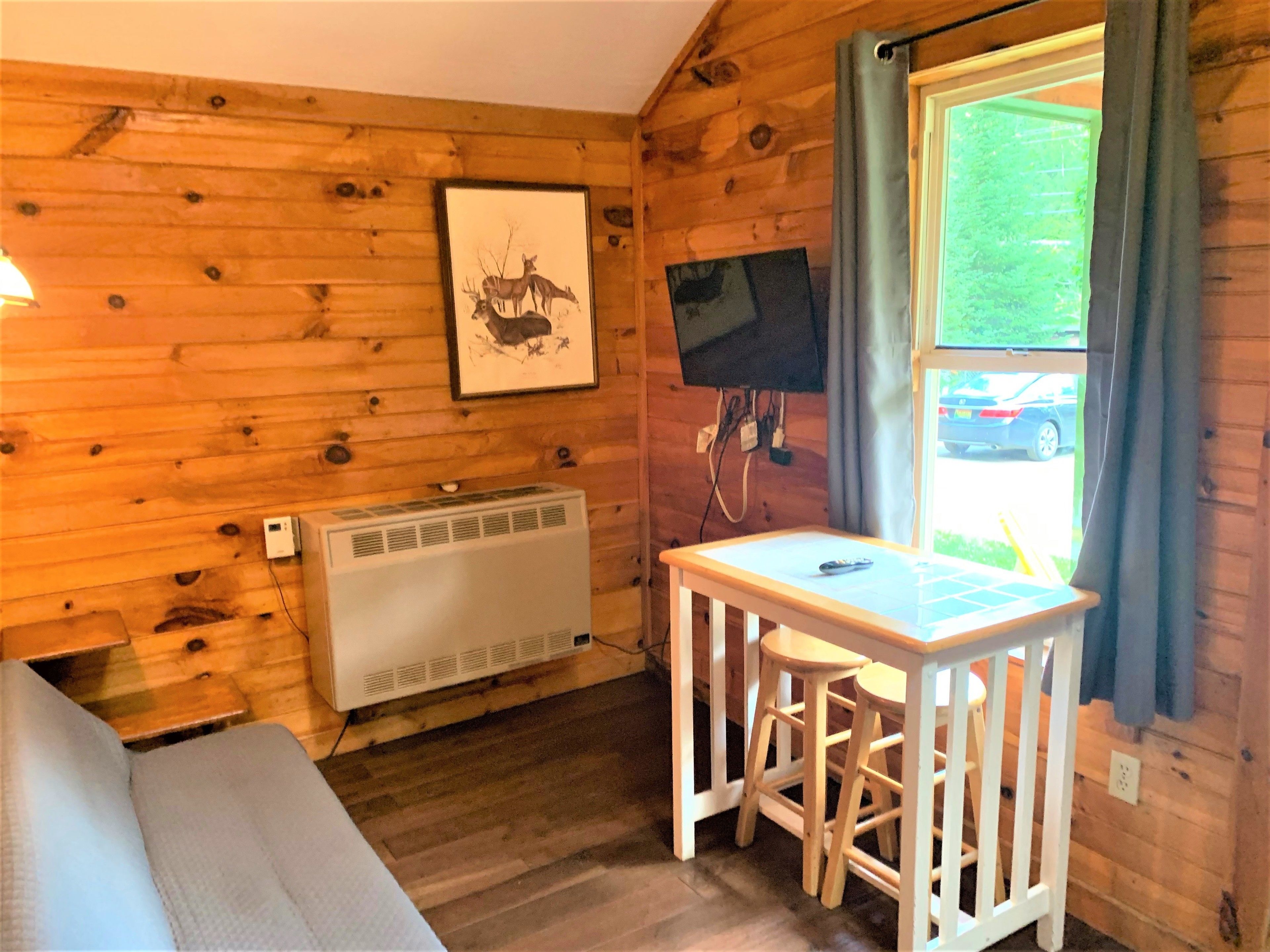 Una habitación de cabaña de madera con una cama, una mesa con dos taburetes, un televisor montado y un cuadro en la pared.