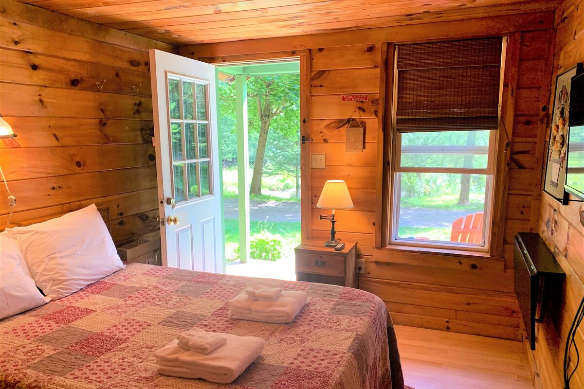 A wooden cabin bedroom with a bed, a nightstand with a lamp, a window with a view of the outdoors, and a door leading outside.