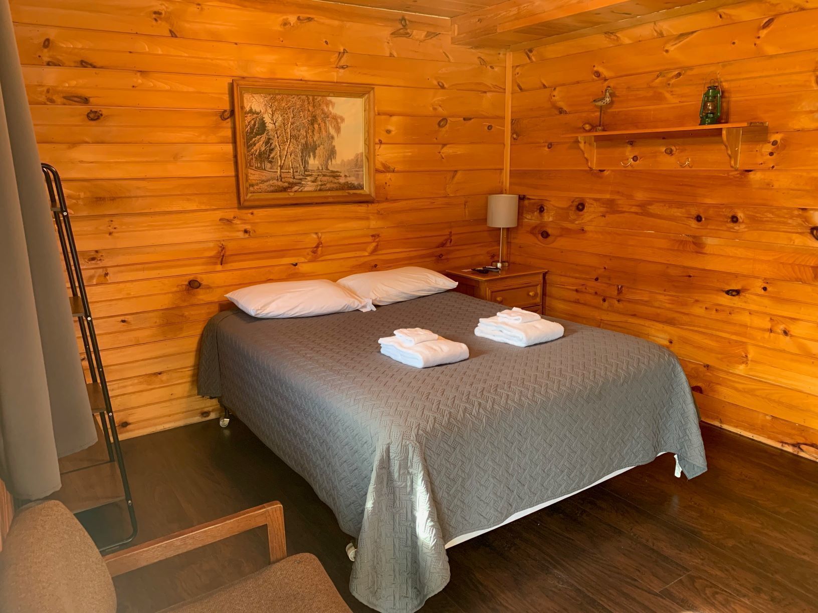 A wooden cabin room with a bed, pillows, a nightstand, a lamp, and a painting on the wall.