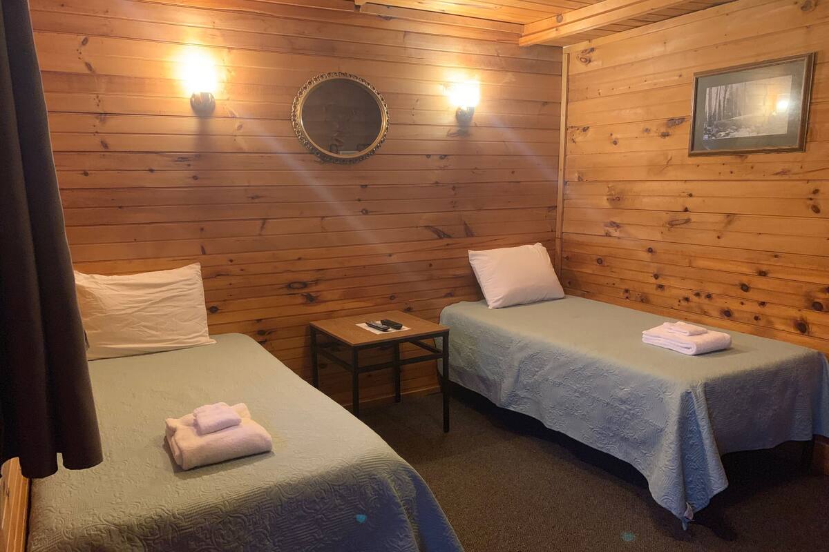 A room with two beds, each with a pillow and a folded towel. There is a small table with a remote control and a framed picture on the wall.