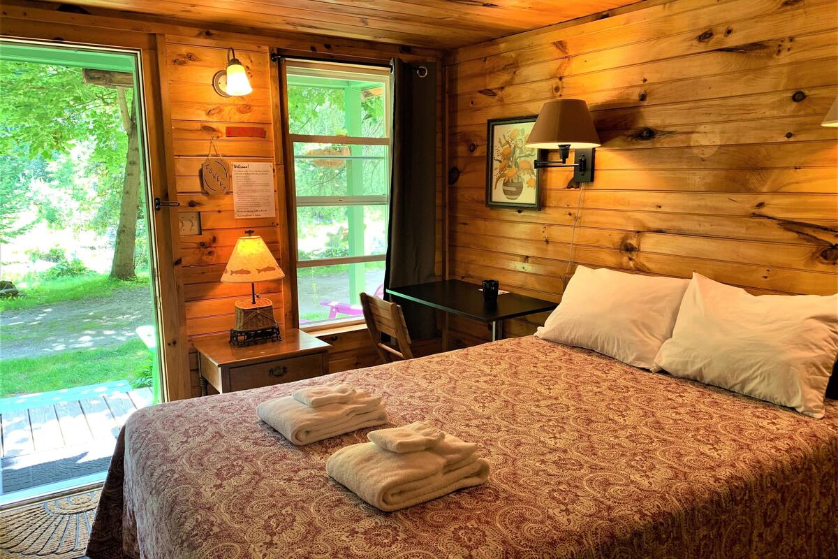 A wooden room with a bed, a table, and a window. There are towels on the bed and a lamp on the table.