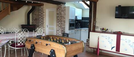 A cozy room with a foosball table, a dining area with a table and chairs, a kitchen with a TV, and a staircase leading to an upper level.