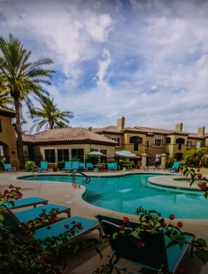 A swimming pool with lounge chairs and umbrellas, surrounded by palm trees and a building with a tiled roof.