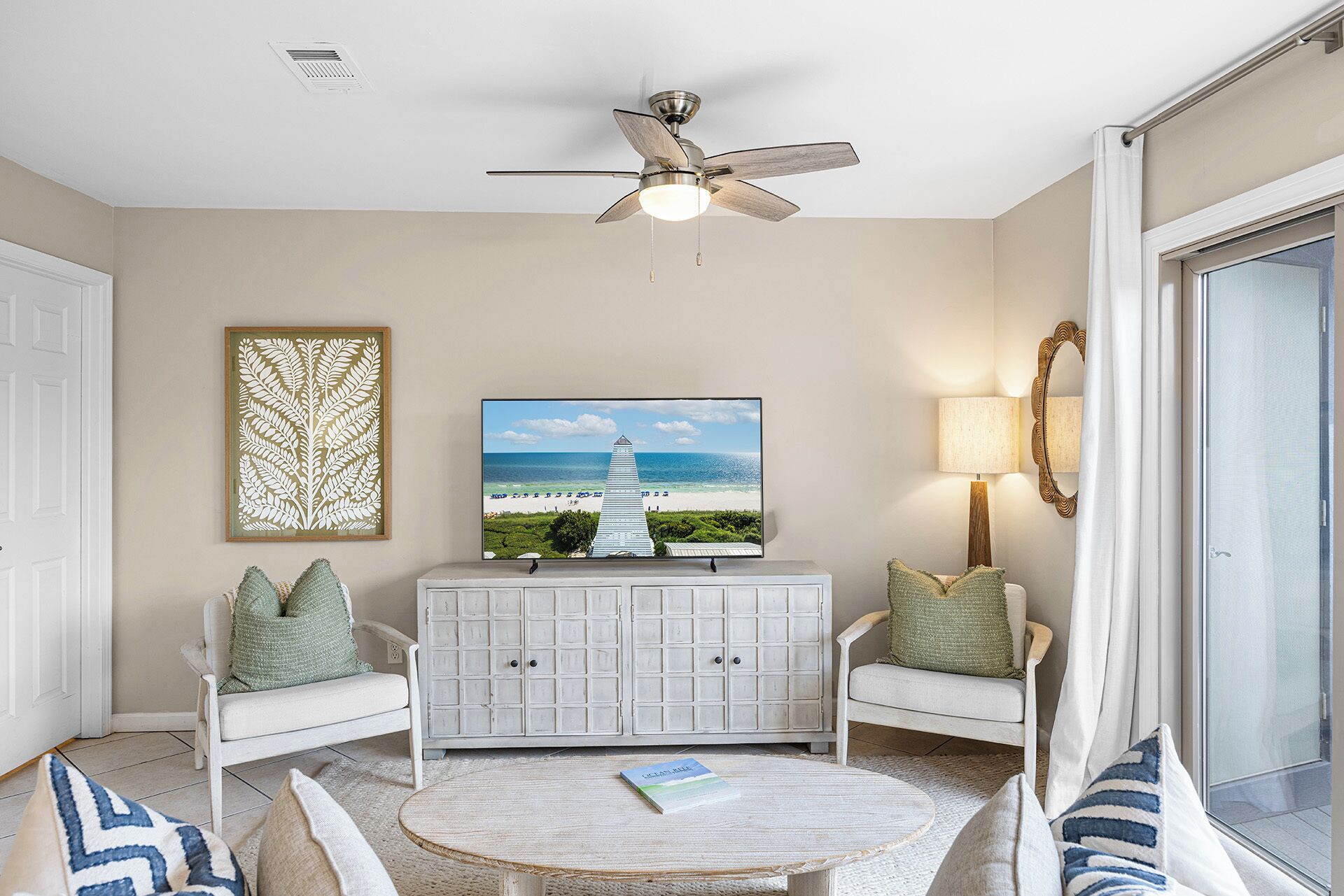 A living room with a ceiling fan, a television displaying a beach scene, a white cabinet, two white armchairs, and a round coffee table.