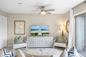 Un salon doté d’un ventilateur de plafond, d’un téléviseur diffusant une scène de plage, d’une armoire blanche, de deux fauteuils blancs et d’une table basse ronde.
