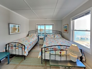 A bedroom with two single beds, a small table, a bench, a window with a view of the sea, and a ceiling fan. - Lindy-B | Sound Views + Screened Porch! (Topsail Beach)