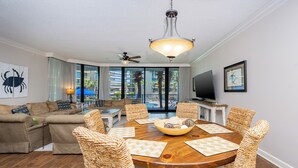 Ein großzügiges Wohnzimmer mit einem runden Holzesstisch, Korbstühlen, einem Deckenventilator und einem großen Fenster mit Blick auf einen Pool und Palmen.