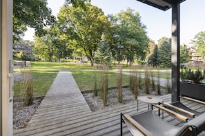 An outdoor deck with a wooden walkway, seating area, and a view of a park with trees and a building in the background. - Uz Juru Mariu (Neringa)