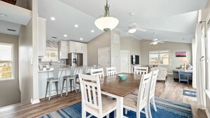 Une cuisine spacieuse avec des meubles blancs, un coin repas avec une table en bois et des chaises blanches, et un salon avec un canapé et un téléviseur.