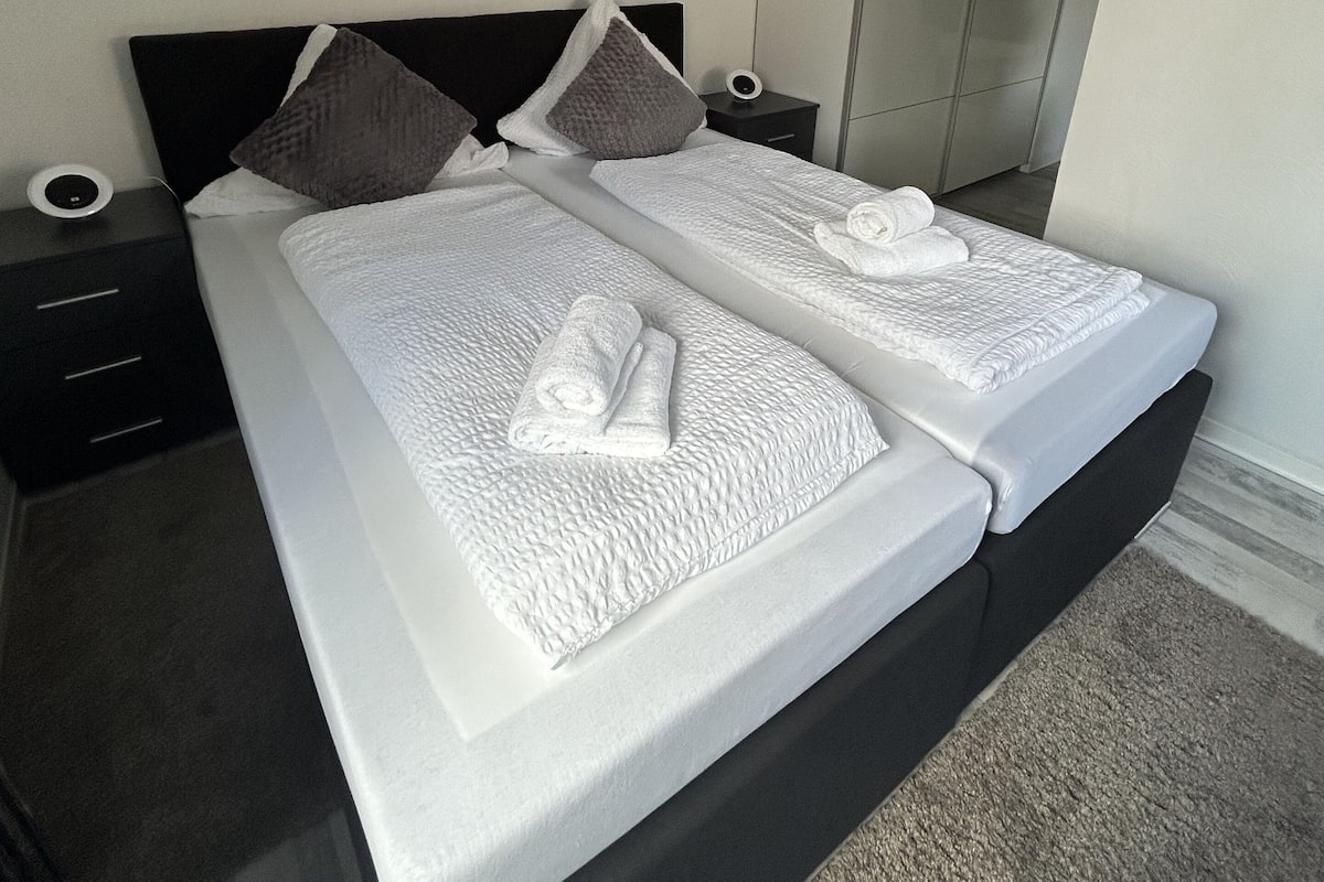 Una cama doble con sábanas blancas y dos almohadas, una mesita de noche con lámpara y un armario al fondo.