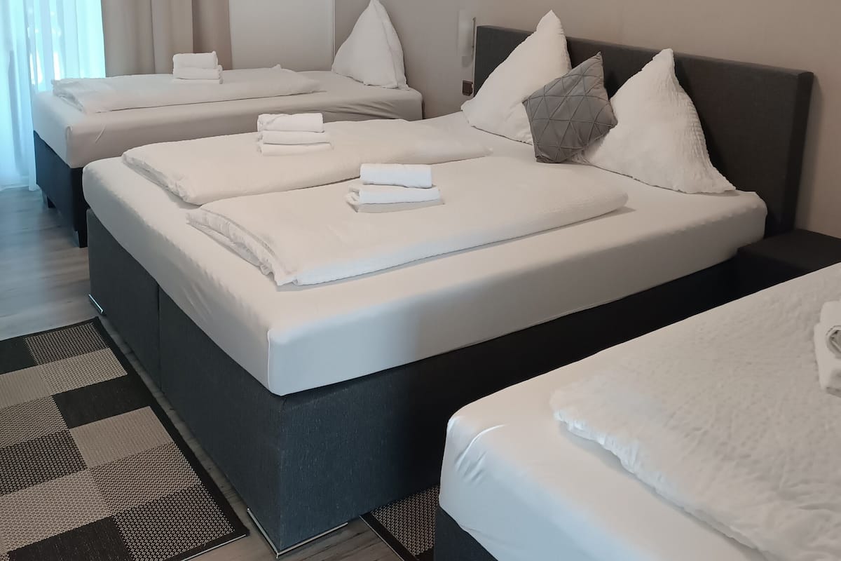 Habitación de hotel con tres camas, cada una con sábanas y almohadas blancas, un equipo de aire acondicionado grande sobre las camas y un mostrador de recepción a la izquierda.