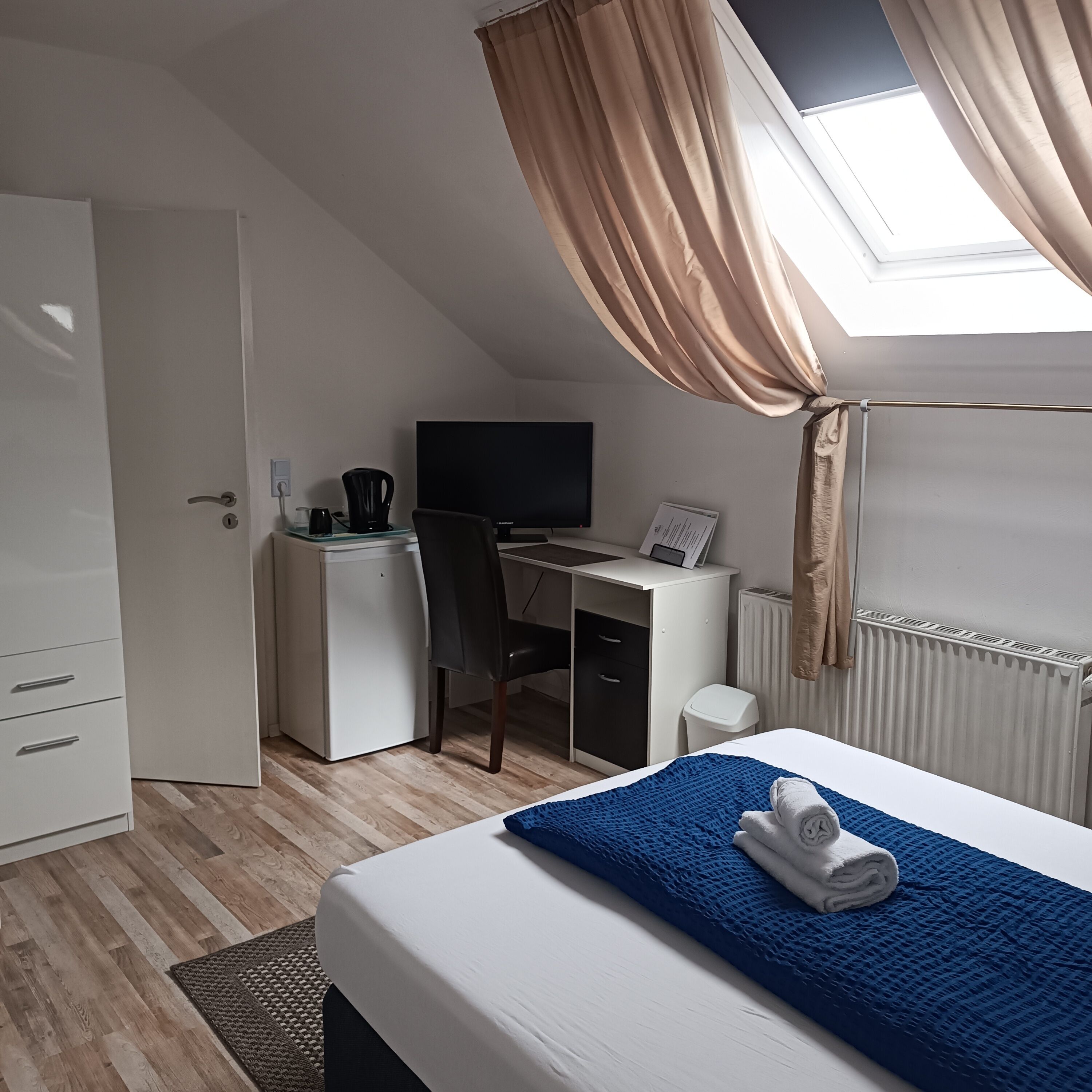 Ein Schlafzimmer mit einem Bett, einem Schreibtisch, einem Stuhl, einem kleinen Kühlschrank, einem Fernseher und einem Fenster mit Vorhängen.