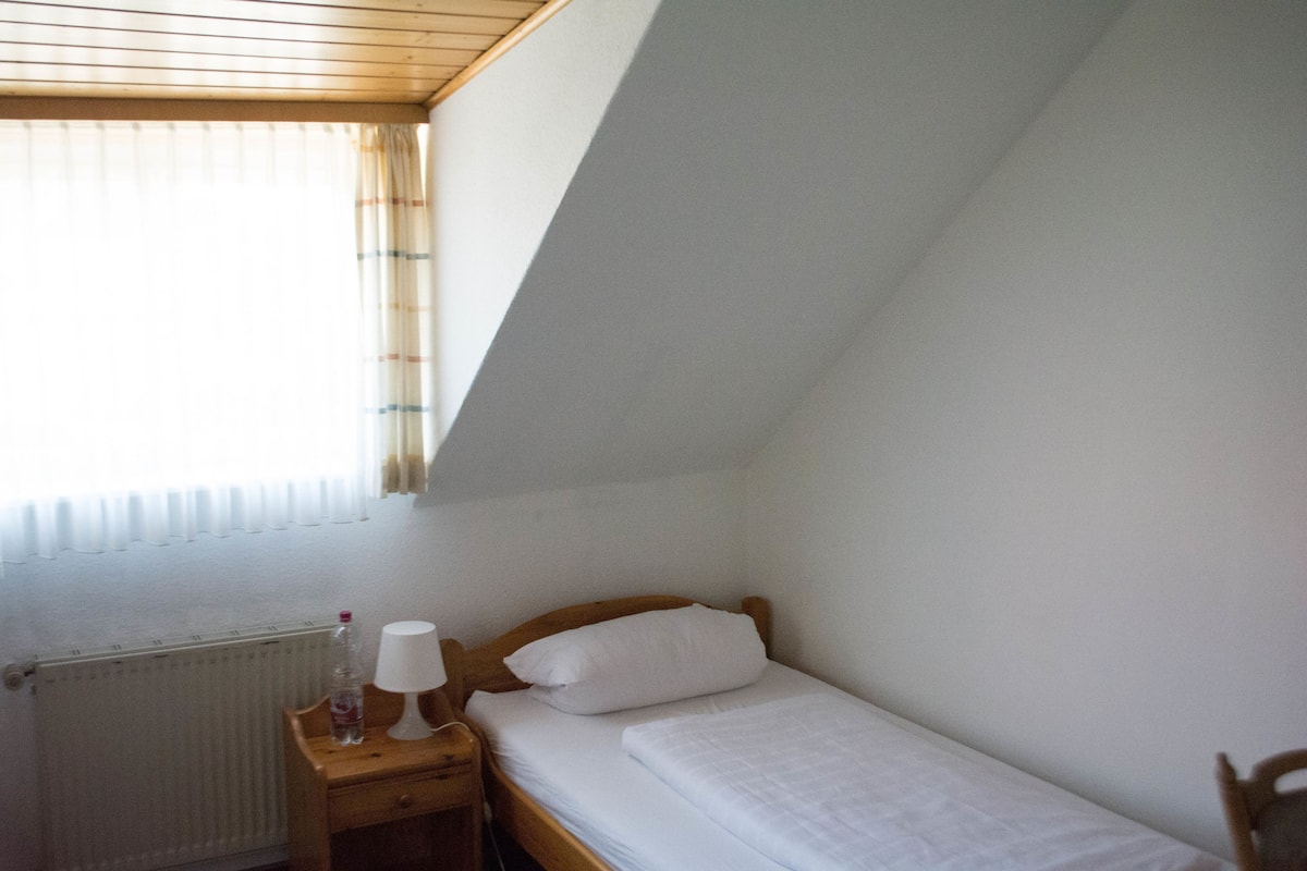 Una habitación pequeña con una cama individual, cabecero de madera, una mesita de noche con lámpara y una ventana con cortinas.
