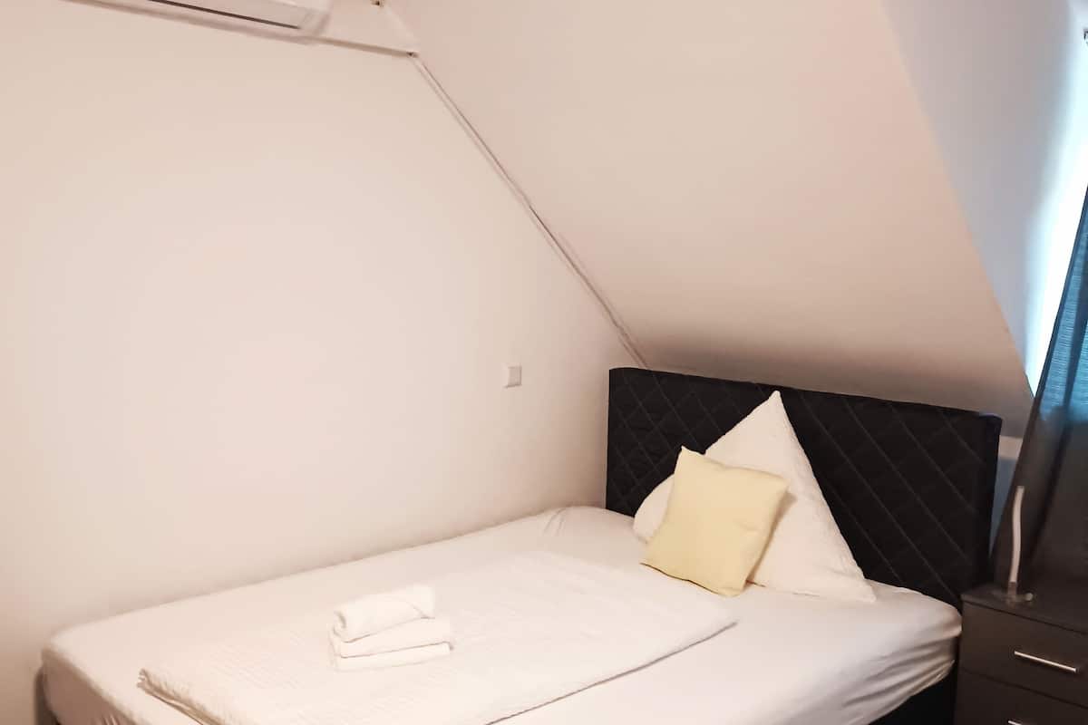 Una cama bien hecha con ropa de cama blanca y una toalla doblada, cabecera oscura y mesita de noche.