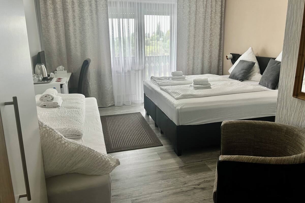 Una habitación de hotel con cama, una silla, un escritorio con tetera y una ventana con cortinas.