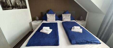 A double bed with blue bedding and two pillows, a nightstand with a lamp, and a painting on the wall.