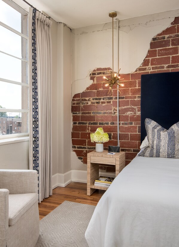 Un dormitorio con pared de ladrillo, mesita de noche de madera, cama, una silla y ventana con cortinas.